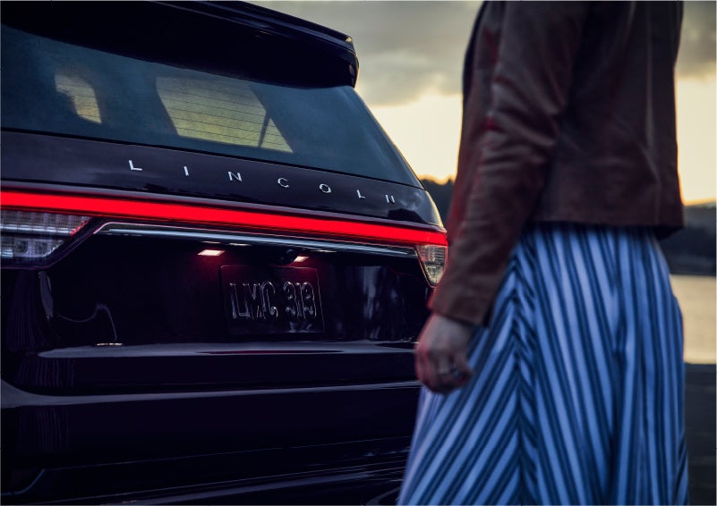 A person is shown near the rear of a 2023 Lincoln Aviator® SUV as the Lincoln Embrace illuminates the rear lights | Green Lincoln in Greensboro NC