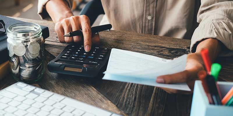 Hands calculating with a calculator, holding documents, with a jar of coins and keyboard on a wooden desk.